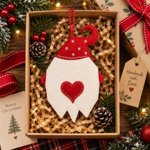 May include: A felt gnome ornament featuring a red hat with white snowflake embroidery, a white beard, and a red heart appliqué. The ornament includes a red hanging loop and is presented in a cardboard gift box filled with crinkle paper.