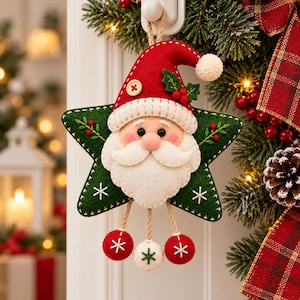 May include: A felt Christmas ornament shaped like a green star with a Santa Claus face in the centre. The Santa wears a red hat with a white pom-pom and a small wooden button. Three small felt balls hang from the bottom on jute strings.