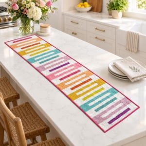 Peut inclure: Un chemin de table matelassé rectangulaire présentant un motif géométrique répétitif de barres parallèles dans un dégradé arc-en-ciel orange, jaune, bleu sarcelle, rose et violet. Le chemin a un fond blanc avec des coutures et une bordure rose.