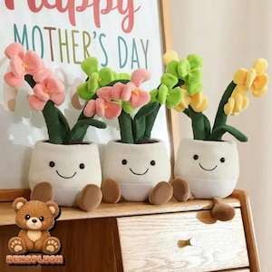 Könnte beinhalten: Drei Plüsch-Blumentöpfe mit lächelnden Gesichtern, die jeweils eine andersfarbige Blume halten: rosa, grün und gelb. Die Töpfe haben braune Füße und stehen auf einer Holzoberfläche. Im Hintergrund steht "Happy Mother's Day".