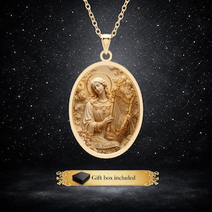May include: A gold-toned oval pendant necklace featuring a relief of Saint Cecilia playing a harp, surrounded by floral details. The pendant hangs from a matching gold chain. Below the pendant, a decorative label reads: Gift box included.