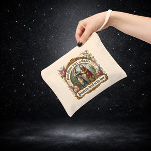 May include: Cream-colored canvas pouch with a wrist strap. The pouch features a framed illustration with the text "Hail Mary Full of Grace" and "Punch the devil in the face." The illustration depicts a woman and a devil-like figure.