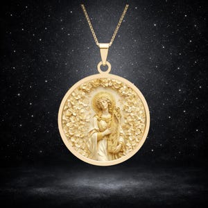 May include: A gold-toned circular pendant necklace featuring a relief of a figure with a halo playing a stringed instrument, surrounded by a dense floral border. The pendant hangs from a delicate gold-toned chain against a dark starry background.