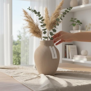 Könnte beinhalten: Eine beige geriffelte Vase mit getrocknetem Pampasgras und Eukalyptuszweigen. Die Vase steht auf einem Holztisch mit einem beigen Leinenläufer. Ein Etikett mit Text hängt an der Vase. Der Raum ist hell und luftig.