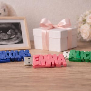 Könnte beinhalten: USB-Sticks mit den Namen "Nicolas" in Blau, "Emma" in Rosa und "Alice" in Grün. Im Hintergrund ein gerahmtes Ultraschallbild, eine weiße Geschenkbox mit rosa Schleife und ein Strauß weißer Rosen.