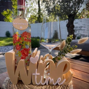 May include: A clear wine bottle with red floral designs and a tag that says "Weinchen?" sits in a wooden holder with a "Wine Time" sign. A wine glass and decorative greenery are also present, all on a wooden table.
