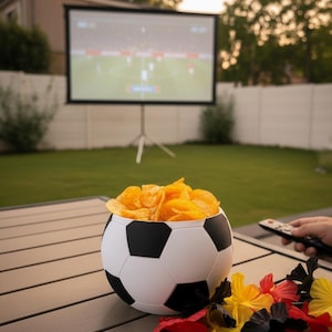 Könnte beinhalten: Eine runde Schüssel im Design eines Fußballs, gefüllt mit goldenen Kartoffelchips, auf einem Lattentisch neben einem kleinen Haufen roter, gelber und schwarzer Stoffblumen, mit einer unscharfen Außenleinwand im Hintergrund.