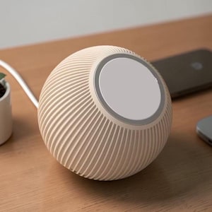 Peut inclure: Un chargeur sans fil sphérique beige avec un dessus blanc et un design nervuré. Une petite plante succulente dans un pot blanc et deux appareils électroniques sont sur une surface en bois.