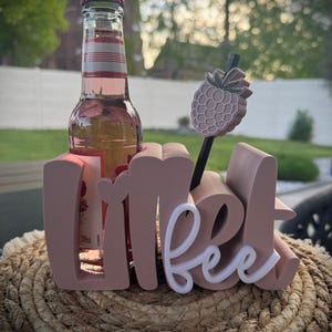 Può includere: Una bottiglia di bevanda rosa in una bottiglia di vetro trasparente, tenuta da un supporto rosa chiaro che recita "sweet bee". Un agitatore per bevande a forma di ananas è nella bottiglia. Il supporto è su un tappetino intrecciato.