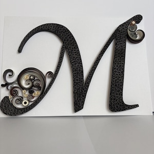 Può includere: Un'opera d'arte di quilling di carta fatta a mano con la lettera "M". La lettera è nera con un motivo a spirale dettagliato. Riccioli decorativi nei toni del nero, marrone e crema accentuano la lettera, su uno sfondo bianco.