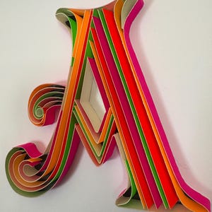 Könnte beinhalten: Ein farbenfrohes Papierkunstwerk in Form des Buchstabens "A". Das Kunstwerk zeigt lebendige Farbtöne von Rosa, Orange, Grün und Rot, wodurch ein dreidimensionaler Effekt entsteht. Der Buchstabe ist vor einem weißen Hintergrund platziert, der die komplexen Papierschichten hervorhebt.
