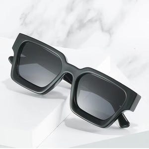 Puede incluir: Gafas de sol negras con montura cuadrada y lentes oscuras. Las gafas de sol tienen una montura gruesa y angular y un dise&ntilde;o moderno. Las lentes son oscuras, proporcionando sombra y protecci&oacute;n contra el sol. Las gafas se muestran sobre una superficie blanca.