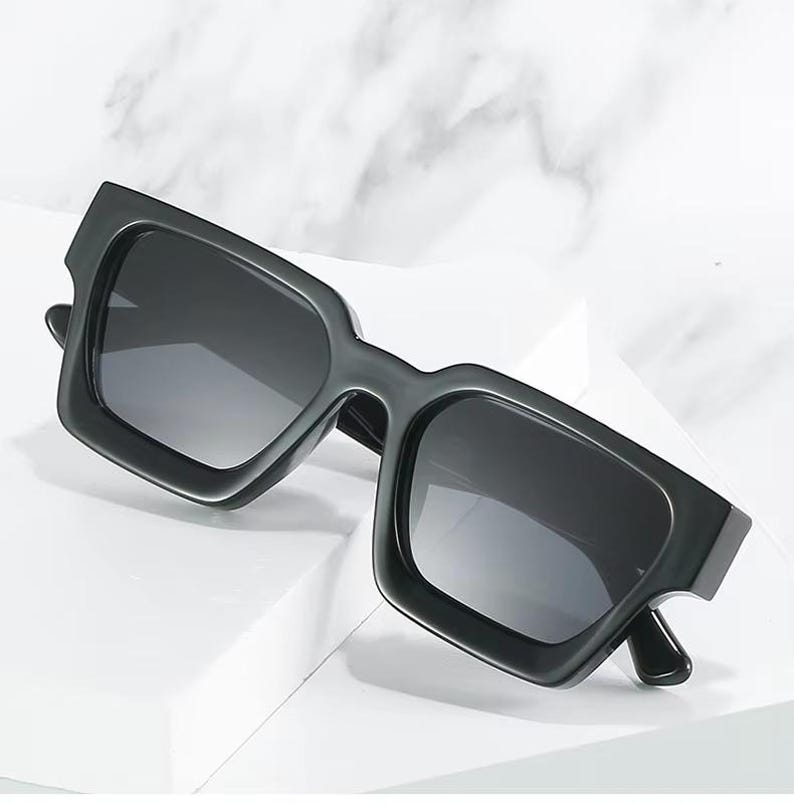 Puede incluir: Gafas de sol negras con montura cuadrada y lentes oscuras. Las gafas de sol tienen una montura gruesa y angular y un dise&ntilde;o moderno. Las lentes son oscuras, proporcionando sombra y protecci&oacute;n contra el sol. Las gafas se muestran sobre una superficie blanca.
