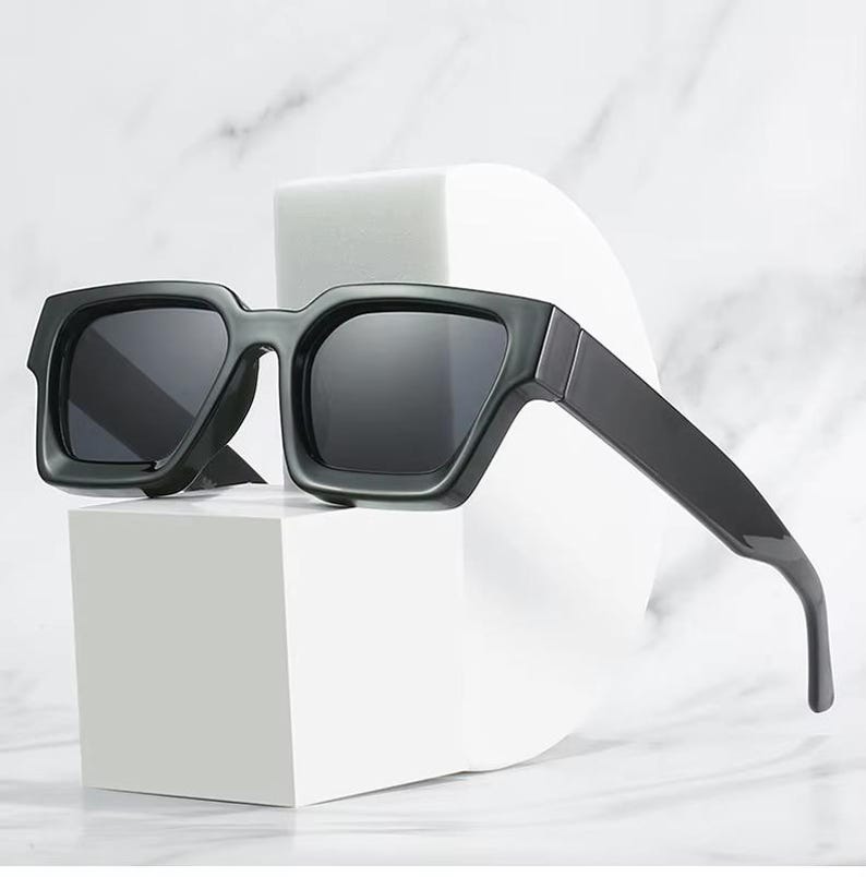 Puede incluir: Gafas de sol grises oscuras con montura cuadrada y lentes oscuras. Las gafas de sol se muestran sobre una forma geom&eacute;trica blanca sobre un fondo de m&aacute;rmol. Las monturas gruesas y el dise&ntilde;o elegante dan un aspecto moderno.