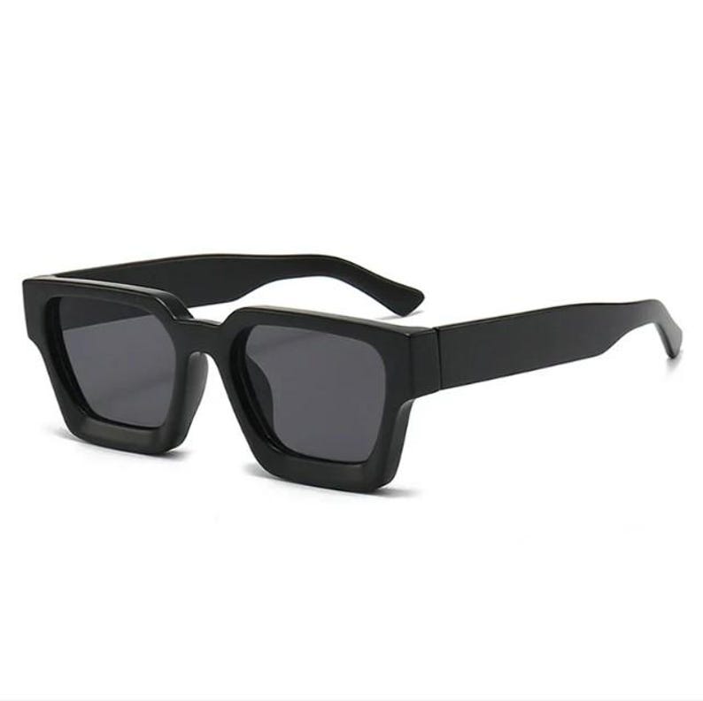 Puede incluir: Gafas de sol cuadradas negras con lentes oscuras. Las gafas tienen una montura y patillas gruesas y atrevidas. Las lentes son de color gris oscuro y la montura es negra lisa. Las gafas son de estilo moderno.