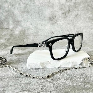 May include: Black eyeglasses with a square frame, silver accents, and a matching chain. The glasses are displayed on a white surface against a gray background. The silver accents feature a cross design.