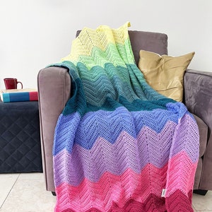 May include: A rainbow chevron crochet throw blanket draped over a gray armchair. The blanket is made of soft yarn and features a gradient of colors from yellow to green, blue, purple, pink, and burgundy.