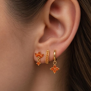 May include: A set of three gold-toned earrings worn in an earlobe. The first is a flower-shaped stud with orange stones, the second is a small hoop encrusted with orange stones, and the third is a gold hoop with a dangling four-petaled orange stone charm.