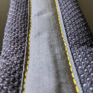 May include: Close-up of a gray fabric item with a textured, woven exterior and a smooth, lighter gray interior. Yellow stitching accents the edges, creating a visual contrast. The item appears to be a strap or binding.