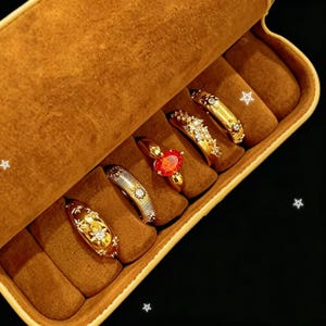 May include: A selection of gold rings presented in a brown jewellery box. The rings showcase diverse designs, including gemstones and detailed patterns. The box is open, displaying the rings in separate compartments.