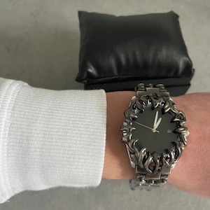 May include: A dark gray wristwatch with a black watch face and a decorative, flame-like bezel. The watch is on a silver-toned metal band. A black leather watch pillow is in the background.
