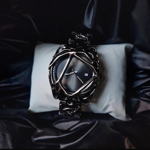 May include: A black wristwatch with a unique, openwork design around the face and band. The watch has a dark gray face with silver hands and a date window. The watch is displayed on a white pillow against a black background.