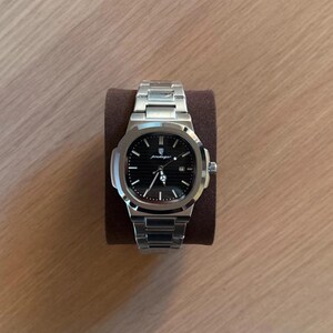 May include: A stainless steel wristwatch with a black dial, featuring a date window and luminous hands. The watch has a silver band and is displayed on a brown watch holder. The watch face has the word "podager" printed on it.
