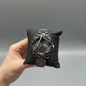 May include: A silver-toned watch with a unique, openwork design around the dark gray face. The watch is displayed on a black cushion, showcasing its intricate details and matching silver band. The watch's design is geometric and artistic.