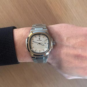 May include: A silver wristwatch with a light-coloured face and a matching metal strap. The watch features a date display and the brand name "podager" is visible. The watch is on a wrist.