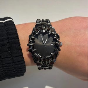 May include: A black wristwatch with a unique, ornate bezel design. The watch face is dark, with silver hands and a date display. The watch is worn on a wrist, with a black sleeve visible in the background. The watch has a bold, masculine aesthetic.