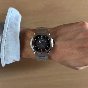 May include: A silver wristwatch with a dark grey face and a silver strap. The watch has a round face with silver hour markers and hands. The watch is on a person's wrist.