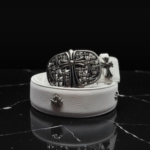 Puede incluir: Cinturón de cuero blanco con hebilla plateada en forma de cruz y adornos en forma de cruz. El cinturón tiene una superficie texturizada y una hebilla decorativa con un diseño de cruz detallado.