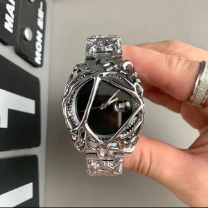 May include: A silver-toned wristwatch with a black face and a unique, textured bezel. The watch features a date display and a matching silver band. The design is intricate and modern, with a focus on geometric shapes and a dark color scheme.