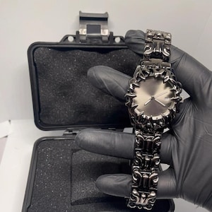 May include: A dark gray wristwatch with a unique, ornate design. The watch features a textured bezel and band, and a light gray watch face. The watch is displayed in an open black case with black foam padding.