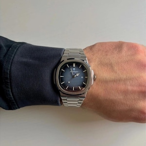 May include: A silver wristwatch with a blue face is displayed on a wrist. The watch features a date window and a stainless steel strap. This is a luxury watch, likely a high-end model.