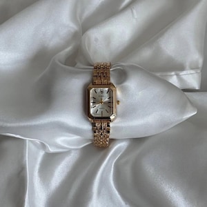 Peut inclure: Une montre-bracelet dorée avec un cadran rectangulaire et un bracelet à maillons assorti. Le cadran de la montre est blanc avec des aiguilles et des marqueurs dorés. La montre est présentée sur un tissu blanc et soyeux.
