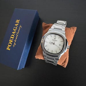 May include: A silver wristwatch with a white face and a date display, presented on a small brown cushion. The watch features a metal strap and is next to a navy blue box with the text "POEDAGAR - top brand luxury".