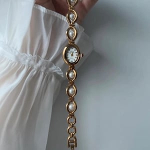 Peut inclure: Montre-bracelet dorée avec un cadran ovale et blanc. La montre est ornée d'un bracelet à maillons avec des fausses perles. La montre est présentée sur un fond blanc, mettant en valeur son design élégant.