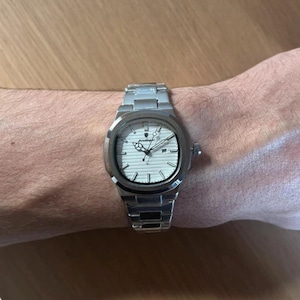 May include: A silver wristwatch with a white face and a silver strap. The watch has a round face with black hands and markers. The watch is on a wrist.