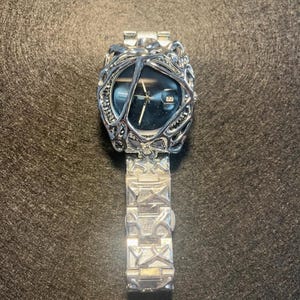May include: A silver-toned wristwatch with an intricate, organic-style bezel and band. The watch face is a deep blue with silver hands and a date display. The band features a repeating geometric pattern.