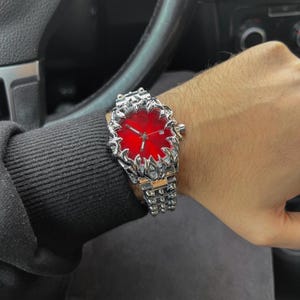 May include: A silver-toned wristwatch with a vibrant red face. The watch features a detailed, textured bezel and band. The watch is worn on a wrist, with a black long-sleeve shirt in the background.