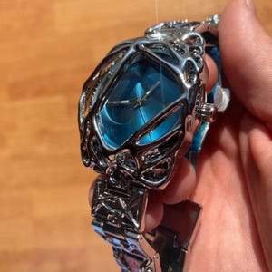 May include: A silver-toned wristwatch with a unique cage-like design around a blue watch face. The watch band is also silver, with an intricate geometric pattern. The watch is held in a hand, showcasing its details.