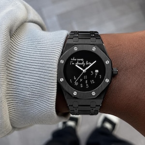 Könnte beinhalten: Eine schwarze Armbanduhr aus Edelstahl mit achteckiger Lünette und sichtbaren Schrauben. Das schwarze Zifferblatt zeigt den weißen Schriftzug 'Who cares I'm already late' und verstreute weiße Zahlen sowie ein Datumsfenster auf der Drei-Uhr-Position.