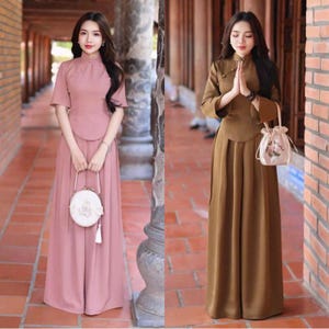 May include: Two women in traditional Asian-style clothing. One wears a pink outfit with a high-necked top and wide-leg trousers, carrying a white purse. The other is in a brown outfit with a similar top and trousers, holding a small bag.