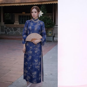 May include: A woman in a navy blue, floral-patterned dress and matching fan. The dress has long sleeves and a high collar. She wears a pearl necklace and a flower in her hair. The background shows a building with a tiled roof and stone walls.