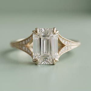 May include: A gold ring featuring a large, rectangular emerald-cut center stone held by four prongs. The band splits into a V-shape on both sides, with each branch encrusted with small, round pavé-set stones that extend along the shank.