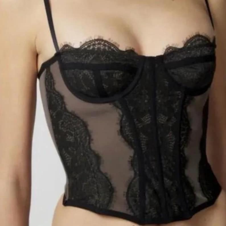 May include: Black lace corset with a sheer beige panel. The corset features delicate lace trim along the top and bottom edges, as well as vertical lace detailing. Thin black straps complete the design.