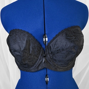 May include: Black strapless bra with a textured, swirling pattern. The bra is displayed on a blue mannequin. The bra has a front closure and a delicate lace trim along the top edge.