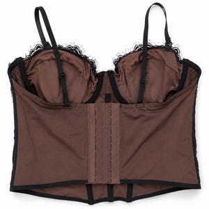 May include: A brown corset with black lace trim and adjustable straps. The corset features a hook-and-eye closure down the front and black straps. The cups are lined with brown fabric and trimmed with black lace.
