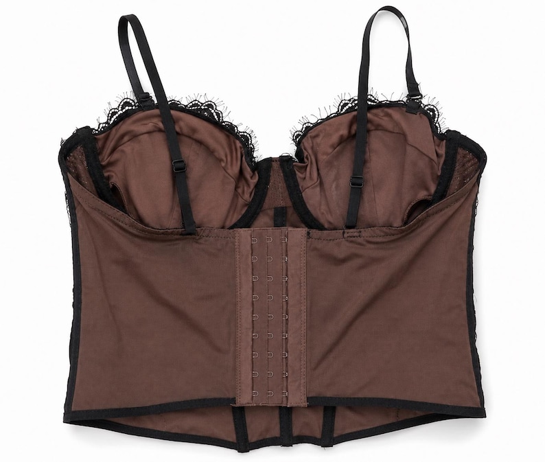 May include: A brown corset with black lace trim and adjustable straps. The corset features a hook-and-eye closure down the front and black straps. The cups are lined with brown fabric and trimmed with black lace.